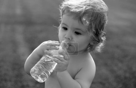Téléchargez les photos : L'enfant boit l'eau d'une bouteille tout en marchant sur le champ d'herbe, la santé du bébé. Eau potable pour bébé - en image libre de droit