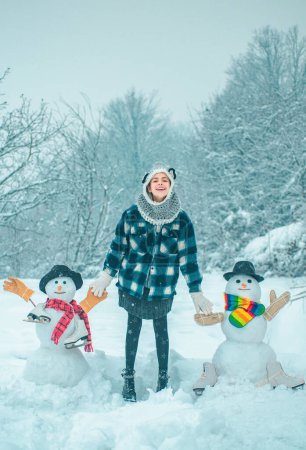 Téléchargez les photos : Scène d'hiver avec des gens heureux sur fond de neige blanche. Le jour d'hiver. Des vêtements de femme d'hiver. Mignon bonhomme de neige dans un village enneigé. Salut bonhomme de neige - en image libre de droit