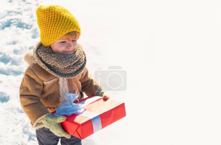 Foto de Niño en ropa de invierno tiene regalo de Navidad. Niño durante el paseo en un parque de invierno nevado. Hermosa naturaleza invernal. Increíble parque de invierno. Adorable niño con cara bonita. Ptoto con espacio de copia vacío - Imagen libre de derechos