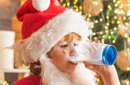 Téléchargez les photos : Père Noël tenant cookie et verre de lait sur fond d'arbre de Noël. Cookies pour l'enfant du Père Noël. Biscuit au lait et pain d'épice pour le Père Noël sur fond bokeh clair de Noël - en image libre de droit