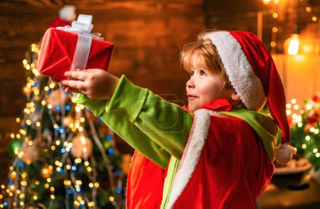 Foto de Niño bastante pequeño en Navidad Santa sombrero tiene una caja de regalo y hermoso fondo decorado de lujo. Feliz Navidad y feliz año nuevo. Niño pequeño con regalos de Navidad - Imagen libre de derechos