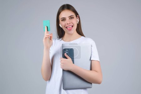 Foto de Mujer joven en camiseta blanca con tarjeta de crédito cerca de la cara y el ordenador portátil, aislado sobre fondo gris. Paga con tarjeta de crédito o débito. Tarjeta bancaria. Tarjeta de crédito de plástico - Imagen libre de derechos