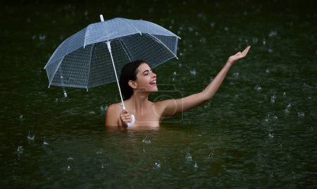 Téléchargez les photos : Femme avec parasol dans l'eau de lac ou de mer. Pluie estivale. Météo pluvieuse. La pluie s'en va. Pluie en été. Femme sexy sensuellement relaxant dans la piscine. Loisirs bien-être - en image libre de droit