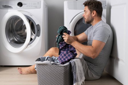 Téléchargez les photos : Homme avec des vêtements près de la machine à laver. Blanchisserie. Ménage, devoirs, femme de ménage. Un mari qui fait la lessive à la maison. Laveuse et sécheuse à la maison - en image libre de droit