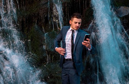 Foto de Empresario con dispositivo en el teletrabajo. Hombre de negocios en traje que sostiene el ordenador portátil en la cascada fondo al aire libre - Imagen libre de derechos