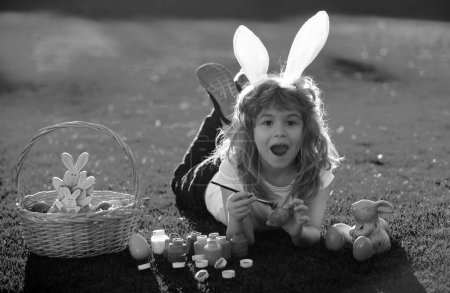 Foto de Niño con huevos de Pascua y orejas de conejo pintando huevos al aire libre. Lindo niño teniendo feliz Pascua en el parque - Imagen libre de derechos