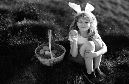Foto de Feliz Pascua para los niños. Niño en orejas de conejo con huevos de colores jugar y la caza de huevos de Pascua fuera. Lindo niño con cesta de Pascua en la hierba - Imagen libre de derechos