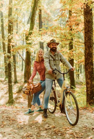 Foto de Feliz pareja hipster sonriente en paseo en bicicleta en el bosque soleado otoñal. Hombre guapo barbudo con sombrero y encantadora mujer atractiva sosteniendo una canasta con hojas de otoño. Concepto de otoño - Imagen libre de derechos