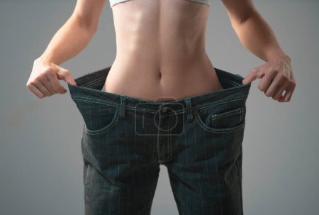 Photo for Skinny waist. Slim girl have points at slim waist in big trousers, successful weight loss, isolated on gray background. Successful weight loss, woman with too large jeans after a diet - Royalty Free Image