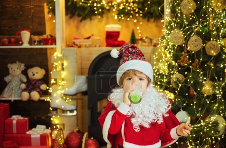 Foto de Niño Santa Claus disfrutando en servido pastel de jengibre y leche. Saludo a la tarjeta de Navidad. Feliz Santa Claus - niño lindo comiendo una galleta y bebiendo un vaso de leche en casa Interior de Navidad - Imagen libre de derechos