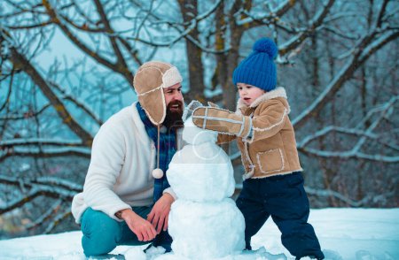 Téléchargez les photos : Père et fils font bonhomme de neige dans la neige. Bonhomme de neige drôle fait main. Vacances de Noël et nouvel an d'hiver avec père et fils. Joyeux famille plaçant avec un bonhomme de neige sur une promenade hivernale enneigée - en image libre de droit