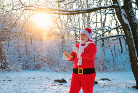 Téléchargez les photos : Père Noël lecture wishlist dans le champ d'hiver forêt enneigée. Père Noël la veille de Noël - en image libre de droit