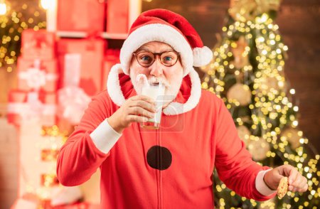 Photo for Portrait of Santa Claus Drinking milk from glass and holding cookies. Santa Claus holding Christmas cookies and milk against Christmas tree background - Royalty Free Image