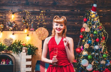 Téléchargez les photos : Belle femme blonde en robe de Noël rouge est debout dans la chambre avec décoration près de l'arbre de Noël brillant et cheminée avec des guirlandes lanternes - en image libre de droit