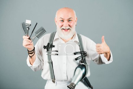 Old happy hairdresser, funny mature barber. Portrait of senior barber holding barber equipments, isolated on grey background. Vintage barbershop, shaving