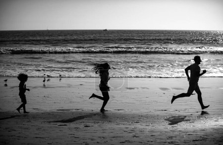 Foto de Siluetas de la familia fitness corriendo cerca del mar. Correr en familia para practicar deporte al aire libre con niños - Imagen libre de derechos