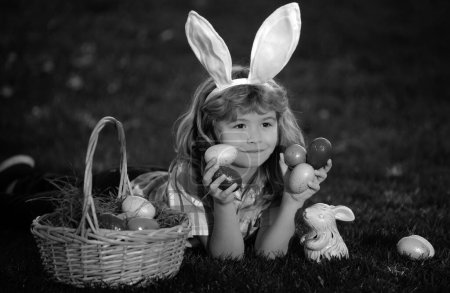Téléchargez les photos : Enfant lapin garçon avec lapin oreilles de lapin. Enfant garçon avec des œufs de Pâques et panier sur l'herbe. Enfant garçon chassant des œufs de Pâques, pondant sur l'herbe. Lapin de Pâques enfants - en image libre de droit