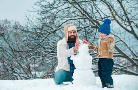 Téléchargez les photos : Fond d'hiver avec flocons de neige et bonhomme de neige. Père et fils font bonhomme de neige dans la neige. Homme de neige drôle fait à la main. Meilleur jeu d'hiver pour une famille heureuse - en image libre de droit