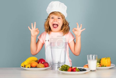 Téléchargez les photos : Chef cuisinier enfant prépare la nourriture sur fond de studio gris isolé. Des enfants cuisinent. Jeune garçon avec tablier et chapeau de chef préparant un légume sain. Chef cuisinier drôle excité - en image libre de droit