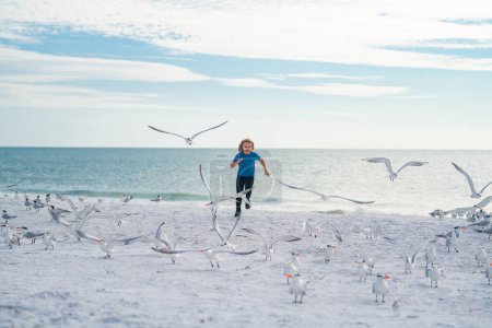 Téléchargez les photos : Vacances d'été. Enfant heureux courant avec les oiseaux de mouettes, s'amusant sur la plage par une chaude journée d'été. Vacances d'été - en image libre de droit