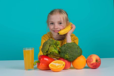 Téléchargez les photos : Fruits et légumes pour enfants. Jolie fille mangeant des fruits frais. Bonne petite fille assise sur la table avec assiette de légumes et de fruits, isolée en studio. Enfant près des légumes et fruits à la cuisine - en image libre de droit
