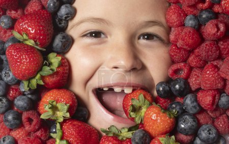 Téléchargez les photos : Vitamines saines fruits. Les enfants font face avec des fruits et des baies. Fraise d'été, myrtille, framboise, fond de mûre. Vue du dessus photo du visage de l'enfant en arrière-plan de baies. Enfants en bonne santé manger - en image libre de droit