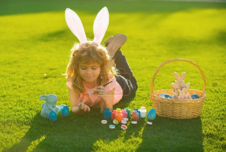 Foto de Niño con traje de conejo con orejas de conejo pintando huevos de Pascua en la hierba en el parque de primavera - Imagen libre de derechos