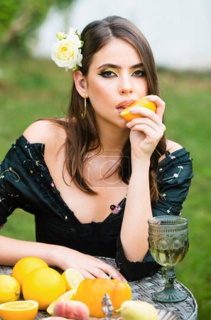 Photo for Sexy woman with red lips, stylish makeup on beautiful face squeezing juice from fresh orange. Outdoor fashion photo of beautiful young woman with rose in hair. Beauty woman eating orange - Royalty Free Image