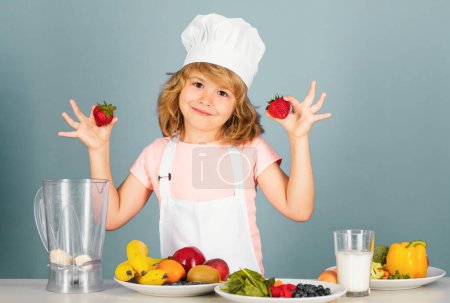 Téléchargez les photos : Chef cuisinier enfant tenir fraises prépare la nourriture sur fond de studio gris isolé. Des enfants cuisinent. Jeune garçon avec tablier et chapeau de chef préparant un repas de légumes sains dans la cuisine - en image libre de droit