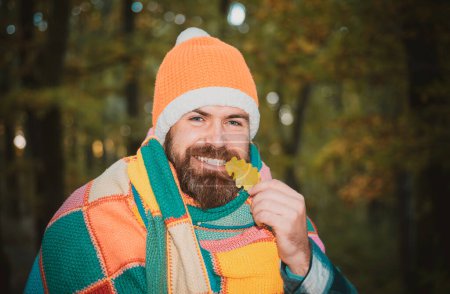 Téléchargez les photos : Hipster gars portant un chapeau orange tricoté et un plaid chaud confortable. Homme heureux d'humeur automnale à l'arrière-plan des arbres. Un homme barbu souriant tient une feuille de chêne automnal dans ses dents - en image libre de droit