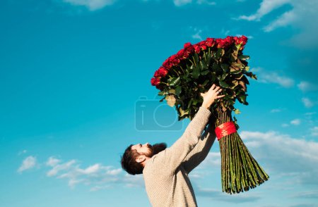 Téléchargez les photos : Homme barbu avec bouquet de roses rouges. Saint Valentin. Cadeau avec amour. Un bel homme sur fond de ciel. Cadeau le jour de la Saint Valentin - en image libre de droit
