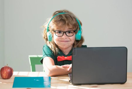 Téléchargez les photos : Un écolier avec des écouteurs qui font des devoirs. Enfant drôle apprend avec ordinateur portable dans la salle d'étude - en image libre de droit