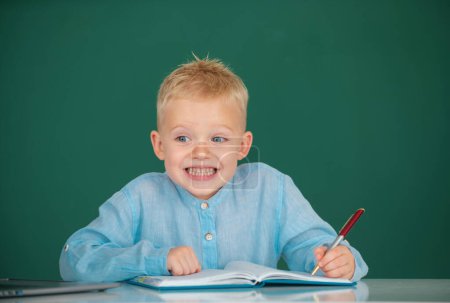 Foto de Cara graciosa de un pequeño estudiante de primaria que estudia en el aula de la escuela. Escribir para niños en cuaderno en clase - Imagen libre de derechos