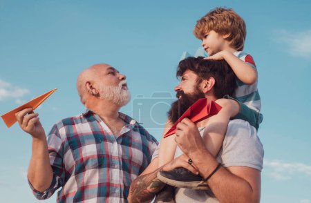 Téléchargez les photos : Heureux grand-père père et petit-fils avec jouet avion en papier sur fond bleu ciel et nuages. Père et fils jouent dehors. Fête des pères. - en image libre de droit