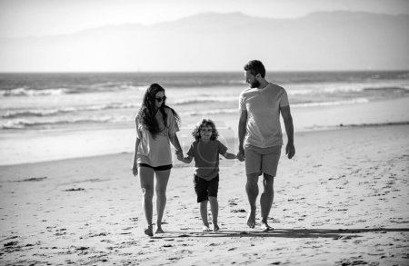 Téléchargez les photos : Heureux père de famille, mère et fils enfant se tiennent la main et marchent le long de la mer surf sur la plage de sable. Parents actifs et personnes en activité de plein air pendant les vacances d'été tropicales avec enfants - en image libre de droit