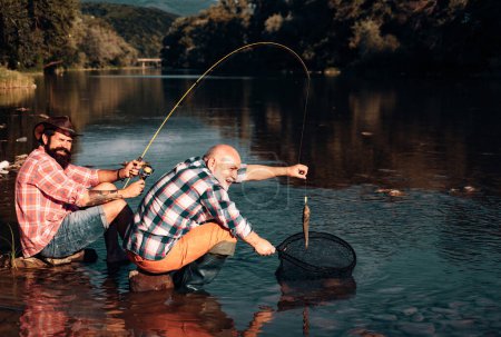 Téléchargez les photos : Attraper des poissons. Portrait d'homme âgé joyeux pêche. Pêcheurs grand-père et fils. Jeune homme et un vieil homme pêchant des filatures sur la rivière ou le lac - en image libre de droit