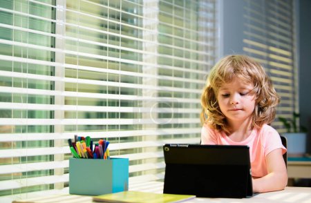 Téléchargez les photos : Petit garçon mignon utilisant une tablette sur le bureau à la maison. Concept d'éducation en ligne. Éducation des enfants, connaissances - en image libre de droit