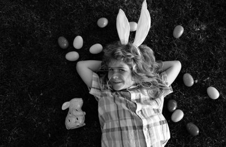 Téléchargez les photos : Lapin de Pâques enfant chasse les œufs en plein air Joyeux jour de Pâques, Garçon couché sur l'herbe dans le parc. Vue de dessus enfants visage avec des oreilles de lapin - en image libre de droit