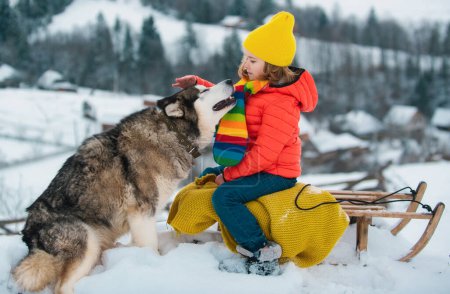 Foto de Niños felices divirtiéndose con perro husky y montando el trineo en el bosque nevado de invierno, disfrutar de la temporada de invierno. Vacaciones de Navidad de invierno y fin de semana de invierno activo, actividades familiares - Imagen libre de derechos