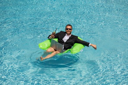 Foto de Un hombre de negocios divertido con un traje de negocios flotando en el agua en la piscina. Trabajo remoto. Freelancer loco. Negocios y verano. Hombre de negocios beber cóctel de verano y el uso de ordenador portátil en la piscina - Imagen libre de derechos