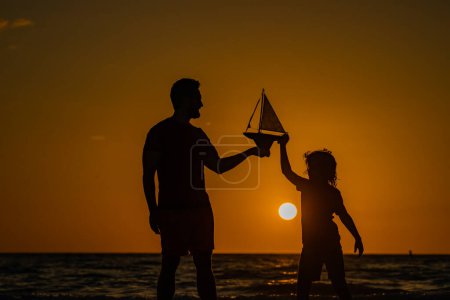 Photo for Father and son playingat the sunset on sea. Father and son play on the sea with toy ship boat on sunrise. Silhouette of father and son. Father and son having fun on sunset beach. Fathers day - Royalty Free Image