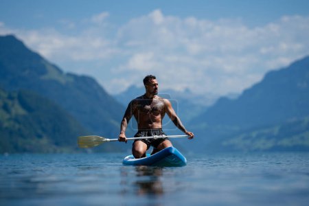 Téléchargez les photos : Homme sur planche à pagaie dans les Alpes montagnes du lac. Loisirs avec pagaie sur le lac en Suisse. Pagaie homme planche de surf sur le lac Léman. Musclé sexy fort hispanique homme pagaie planches surfeurs - en image libre de droit