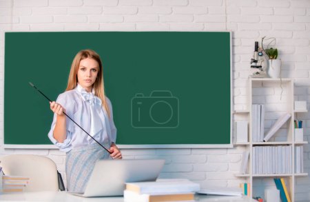 Foto de Joven profesora rubia señalando la lección. Linda mujer joven con puntero enseñanza cerca de pizarra - Imagen libre de derechos