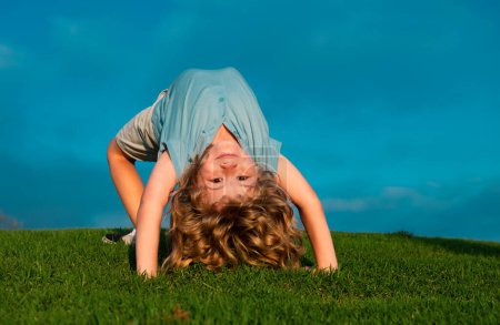 Téléchargez les photos : Enfant garçon debout à l'envers sur l'herbe verte dans le parc d'été. Le concept d'un mode de vie familial sain. L'enfance amusante et insouciante. Sport pour enfants sur la nature - en image libre de droit