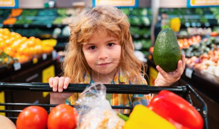 Foto de El niño está de compras en un supermercado. Comida saludable para familias jóvenes con niños. Retrato de niño sonriente con carrito de compras lleno de verduras frescas. Niños en la tienda de comestibles o supermercado - Imagen libre de derechos