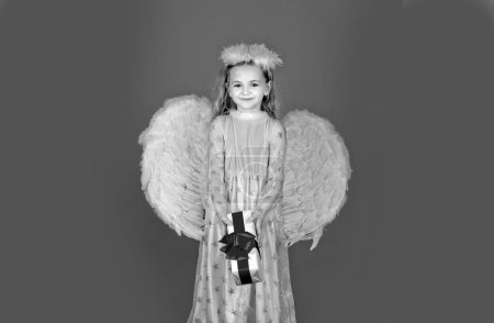 Téléchargez les photos : Petit ange enfant avec cadeau. Petit ange aux ailes blanches tient cadeau. Enfant ange mignon avec des ailes d'anges, isolé sur rouge. Carte cadeau Saint Valentin - en image libre de droit