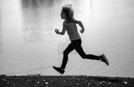 Téléchargez les photos : Enfants courir ou faire du jogging près du lac sur l'herbe dans le parc. Enfant sportif garçon coureur courir dans le parc d'été. Des enfants actifs, des enfants sportifs. Course matinale avec enfants - en image libre de droit