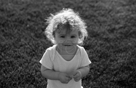 Foto de Bebé feliz en la hierba en el fieald en la soleada noche de verano. Sonriendo niño al aire libre. Primer plano de la cara de bebé. Gracioso niño pequeño cerca de retrato. Niño rubio, cara de emoción sonriente - Imagen libre de derechos