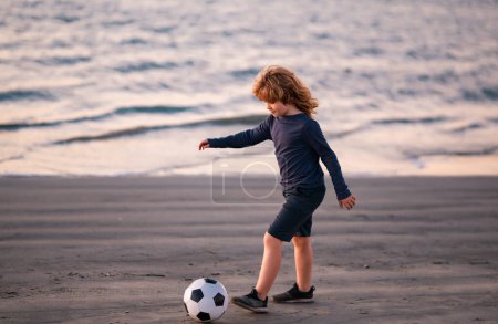 Photo for Young soccer player in sportswear with soccer ball. Cheerful little boy enjoy soccer, football sport games on sunset beach - Royalty Free Image