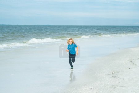 Foto de El chico corre en la playa de verano. Niño corriendo a lo largo del océano. El niño corre a lo largo de la orilla del mar en las vacaciones de verano. Feliz niño descansa en el mar. Concepto vacaciones de verano - Imagen libre de derechos